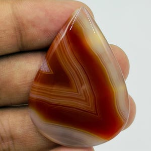 Pode incluir: Uma pedra preciosa de &aacute;gata polida em forma de l&aacute;grima com faixas conc&ecirc;ntricas de laranja queimado profundo, castanho avermelhado e branco creme transl&uacute;cido. A pedra tem uma superf&iacute;cie lisa e brilhante com um padr&atilde;o de chevron vis&iacute;vel.