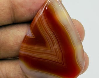 May include: A teardrop-shaped polished agate gemstone featuring concentric bands of deep burnt orange, reddish-brown, and translucent cream white. The stone has a smooth, glossy surface with a distinct chevron pattern visible within the layered mineral structure.
