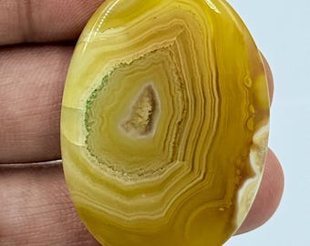 Puede incluir: Una piedra preciosa de ágata amarilla de forma ovalada con patrones de bandas concéntricas en varios tonos de amarillo mostaza y dorado, con un centro cristalino texturizado y un acabado de superficie pulido.
