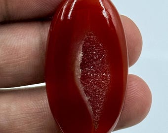 Puede incluir: Una piedra preciosa de cornalina pulida de forma ovalada, con una superficie lisa de color rojo intenso y una fisura central vertical que revela un interior druzy cristalino y texturizado que brilla con pequeños depósitos minerales claros.