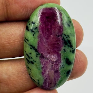Op de afbeelding: Een ovaalvormige cabochon edelsteen met een levendig magenta-roze centrum omringd door lichtgroene zoisiet en verspreide zwarte hoornblende insluitingen. Het gepolijste oppervlak heeft een gladde, glanzende afwerking met een duidelijk contrast.