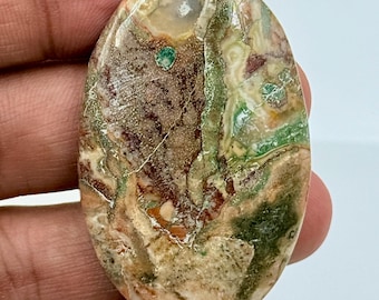 May include: An oval-shaped polished gemstone cabochon featuring a complex pattern of earthy brown, cream, and moss green tones. The surface displays intricate natural inclusions, swirling textures, and small translucent spots throughout the stone.