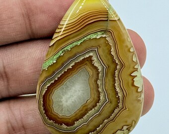 Puede incluir: Una piedra preciosa de ágata pulida en forma de lágrima con bandas concéntricas de colores amarillo, marrón y crema, un núcleo central de cristal blanco translúcido y una fina veta de inclusión mineral verde.