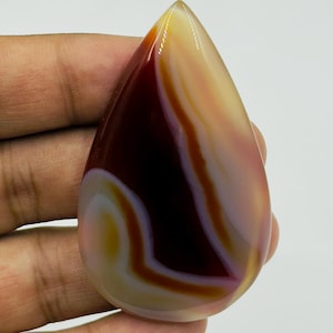 May include: A polished, teardrop-shaped agate gemstone featuring smooth, flowing bands of deep maroon, translucent cream, and soft pink. The stone has a glossy, reflective surface with distinct, organic color transitions and a pointed top.