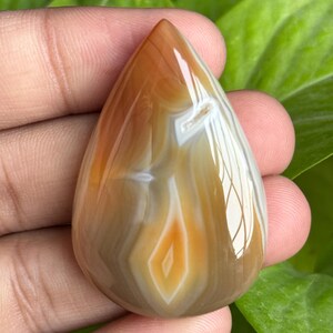 May include: A polished, teardrop-shaped gemstone with a warm color palette. The stone features bands of brown, white, and orange hues, creating a natural, layered pattern. A central diamond-shaped design adds a focal point to the polished surface.