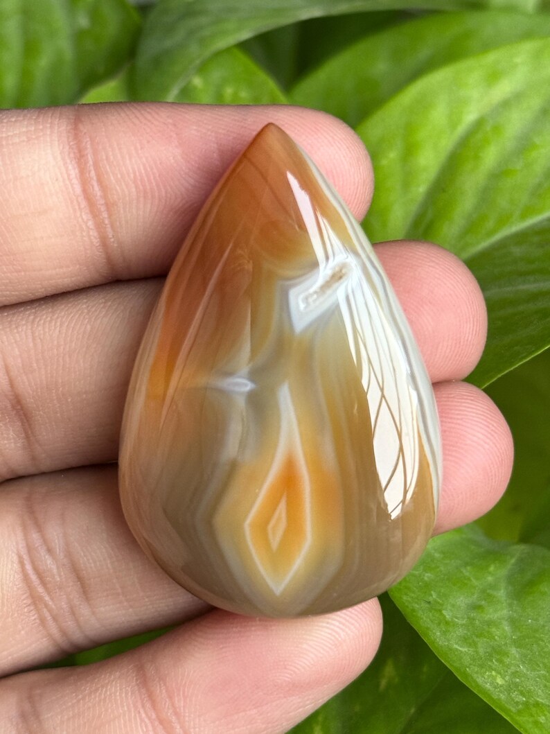 May include: A polished, teardrop-shaped gemstone with a warm color palette. The stone features bands of brown, white, and orange hues, creating a natural, layered pattern. A central diamond-shaped design adds a focal point to the polished surface.