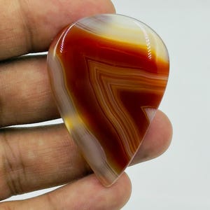 Pode incluir: Uma pedra preciosa de &aacute;gata polida em forma de l&aacute;grima com faixas vibrantes de laranja queimado profundo, vermelho ferrugem e branco transl&uacute;cido. A pedra tem uma superf&iacute;cie lisa e brilhante com padr&otilde;es geom&eacute;tricos n&iacute;tidos e definidos.