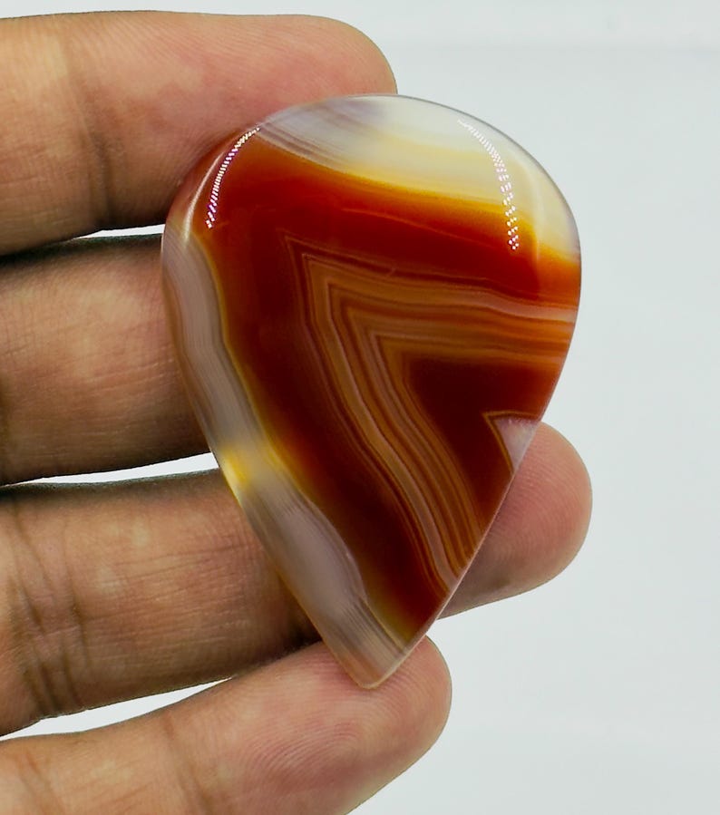 Pode incluir: Uma pedra preciosa de &aacute;gata polida em forma de l&aacute;grima com faixas vibrantes de laranja queimado profundo, vermelho ferrugem e branco transl&uacute;cido. A pedra tem uma superf&iacute;cie lisa e brilhante com padr&otilde;es geom&eacute;tricos n&iacute;tidos e definidos.