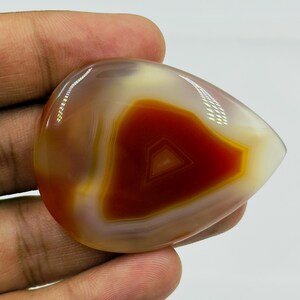 May include: A polished, teardrop-shaped agate gemstone featuring concentric bands of deep burnt orange, translucent amber, and creamy white. The stone has a smooth, glossy surface with visible light reflections across its curved, rounded edges.