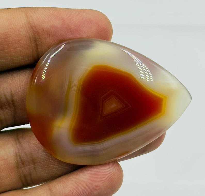 May include: A polished, teardrop-shaped agate gemstone featuring concentric bands of deep burnt orange, translucent amber, and creamy white. The stone has a smooth, glossy surface with visible light reflections across its curved, rounded edges.