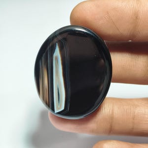 May include: A polished, oval-shaped gemstone with a black base and layered bands of white, tan, and light blue. The stone is held in a hand, showcasing its smooth, reflective surface. The gemstone's design is a natural, organic pattern.