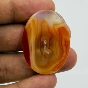 May include: An oval-shaped polished agate gemstone featuring translucent layers of orange, amber, and cream colors. The stone displays natural concentric banding patterns with a central white inclusion and a smooth, glossy surface finish.