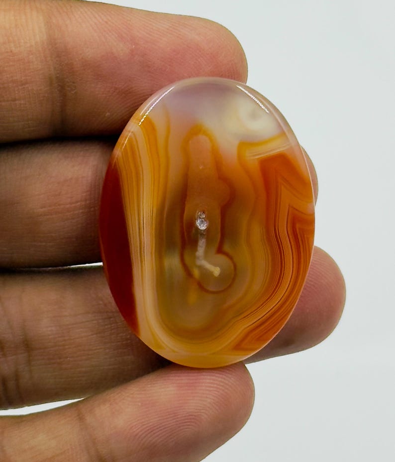 May include: An oval-shaped polished agate gemstone featuring translucent layers of orange, amber, and cream colors. The stone displays natural concentric banding patterns with a central white inclusion and a smooth, glossy surface finish.