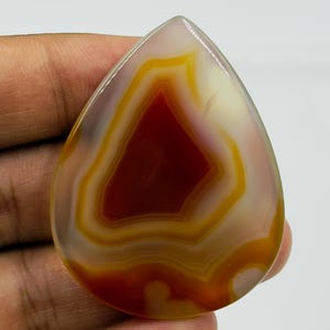 May include: A polished, teardrop-shaped agate gemstone featuring concentric bands of deep red, burnt orange, and translucent white. The stone has a smooth, glossy surface with a distinct layered pattern radiating from a dark red center.