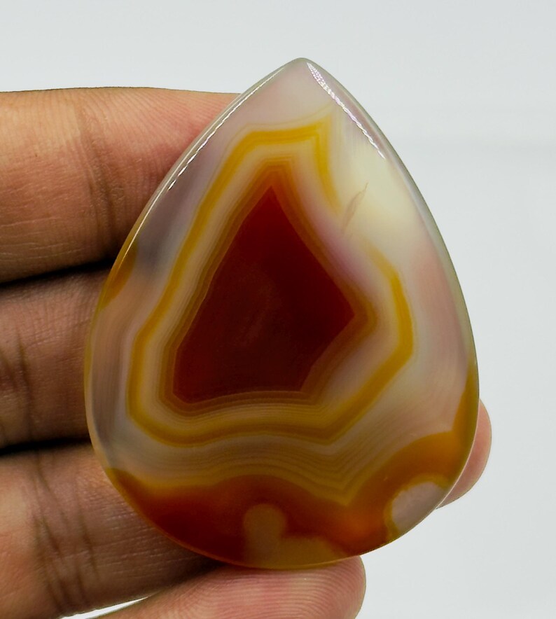 May include: A polished, teardrop-shaped agate gemstone featuring concentric bands of deep red, burnt orange, and translucent white. The stone has a smooth, glossy surface with a distinct layered pattern radiating from a dark red center.