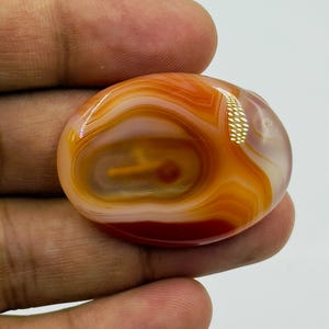 May include: An oval-shaped cabochon gemstone featuring concentric bands of translucent orange, white, and deep red colors. The polished stone displays a smooth, glossy surface with natural swirling patterns and a distinct, centered circular inclusion.