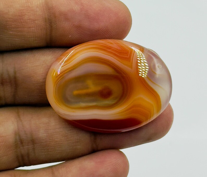 May include: An oval-shaped cabochon gemstone featuring concentric bands of translucent orange, white, and deep red colors. The polished stone displays a smooth, glossy surface with natural swirling patterns and a distinct, centered circular inclusion.
