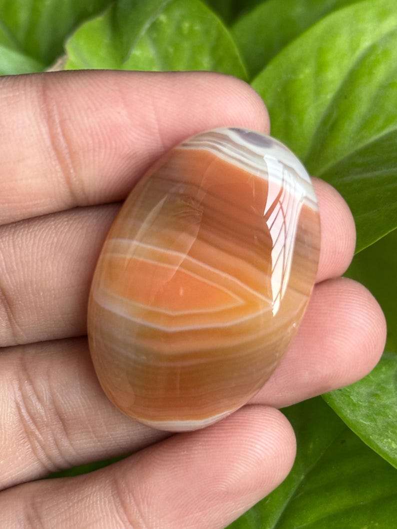 May include: Oval-shaped polished gemstone with layers of orange, brown, and white bands. The stone has a glossy surface and is held in a hand against a backdrop of green leaves. This gemstone is likely used for jewelry or decorative purposes.