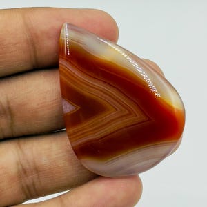 Pode incluir: Uma pedra preciosa de &aacute;gata polida em forma de l&aacute;grima com faixas vibrantes de laranja profundo, vermelho queimado e branco transl&uacute;cido. A pedra exibe uma superf&iacute;cie lisa e brilhante com padr&otilde;es angulares conc&ecirc;ntricos e distintos.