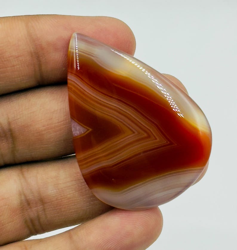 Pode incluir: Uma pedra preciosa de &aacute;gata polida em forma de l&aacute;grima com faixas vibrantes de laranja profundo, vermelho queimado e branco transl&uacute;cido. A pedra exibe uma superf&iacute;cie lisa e brilhante com padr&otilde;es angulares conc&ecirc;ntricos e distintos.