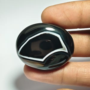 May include: A polished, oval-shaped gemstone with a striking black and white banded pattern. The stone features a central black area surrounded by a white, angular design, creating a bold contrast. The gem is held between fingers, showcasing its smooth, reflective surface.