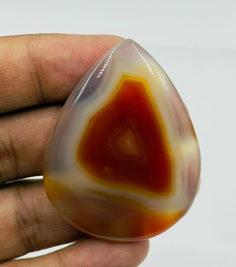 May include: A polished, teardrop-shaped agate gemstone featuring concentric bands of deep burnt orange, translucent white, and soft cream colors. The stone has a smooth, glossy surface with a distinct triangular pattern visible in the center.