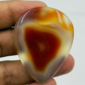 May include: A polished, teardrop-shaped agate gemstone featuring concentric bands of deep rust-red, warm amber, and translucent cream colors. The stone has a smooth, glossy surface with a distinct geometric triangular pattern visible in the center.