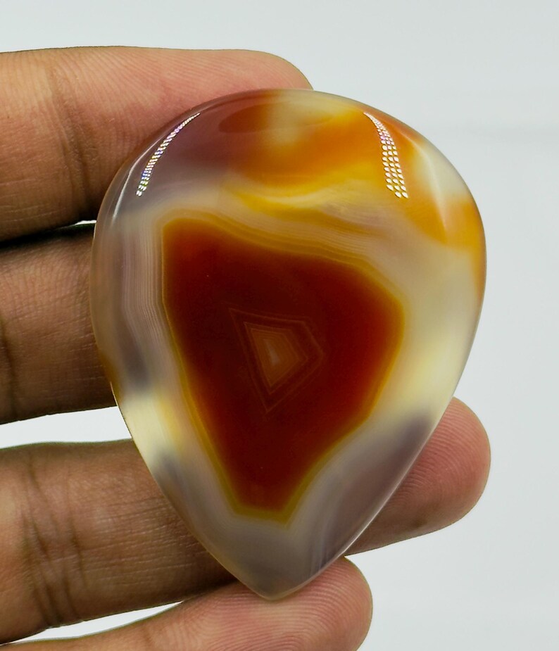 May include: A polished, teardrop-shaped agate gemstone featuring concentric bands of deep rust-red, warm amber, and translucent cream colors. The stone has a smooth, glossy surface with a distinct geometric triangular pattern visible in the center.