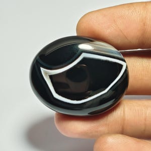 May include: A polished, round gemstone with a striking black and white banded pattern. The stone features a prominent white, eye-like design against a glossy black background. The gemstone is held between fingers, showcasing its smooth, reflective surface.
