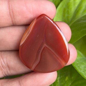 May include: A polished, teardrop-shaped gemstone in shades of red and brown, with white veining. The stone is held against a backdrop of green foliage, highlighting its natural beauty and unique patterns. This gemstone is ideal for jewelry making.