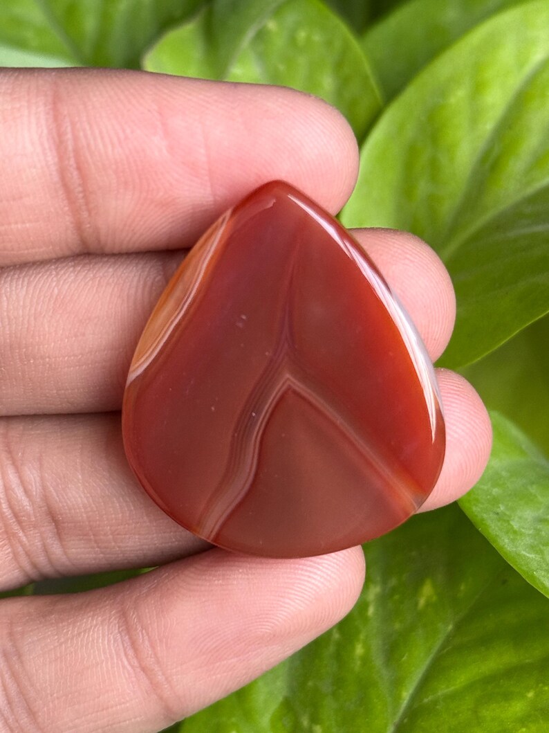 May include: A polished, teardrop-shaped gemstone in shades of red and brown, with white veining. The stone is held against a backdrop of green foliage, highlighting its natural beauty and unique patterns. This gemstone is ideal for jewelry making.