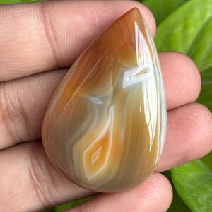May include: A polished, teardrop-shaped agate gemstone. The stone features bands of brown, tan, and white, with a glossy finish. The pattern includes a diamond-shaped inclusion. The gemstone is held in a hand.