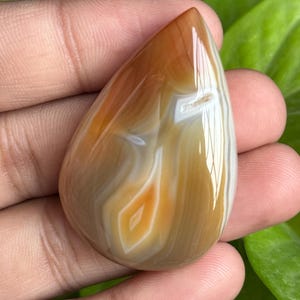 May include: A polished, teardrop-shaped gemstone with a warm color palette. The stone features bands of brown, tan, and white, with a central diamond-shaped pattern in orange and white. The gemstone is held in a hand, showcasing its smooth, reflective surface.