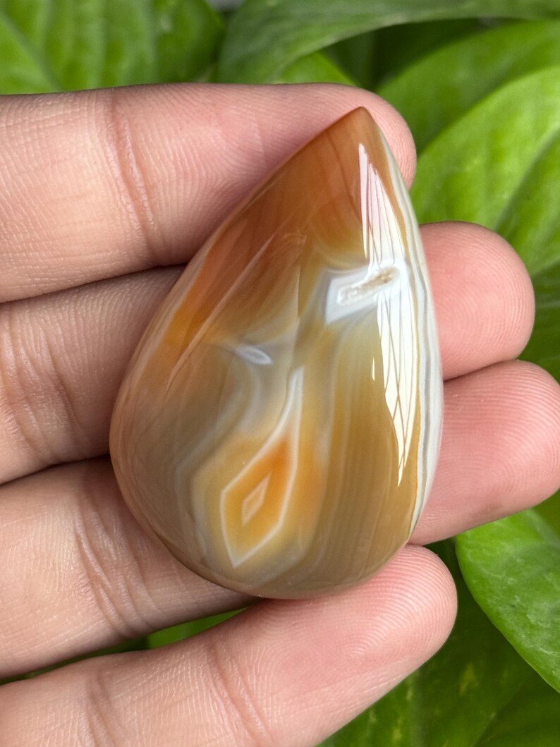 May include: A polished, teardrop-shaped gemstone with a warm color palette. The stone features bands of brown, tan, and white, with a central diamond-shaped pattern in orange and white. The gemstone is held in a hand, showcasing its smooth, reflective surface.