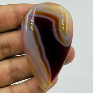 May include: A polished, teardrop-shaped agate gemstone featuring smooth, concentric bands of deep mahogany, translucent cream, and soft lavender. The stone has a glossy, reflective surface with distinct color transitions and a pointed bottom.