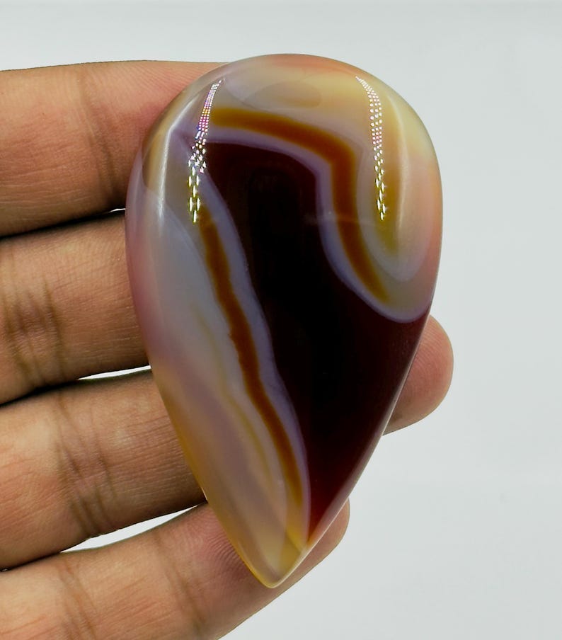 May include: A polished, teardrop-shaped agate gemstone featuring smooth, concentric bands of deep mahogany, translucent cream, and soft lavender. The stone has a glossy, reflective surface with distinct color transitions and a pointed bottom.