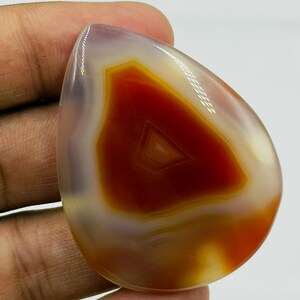 May include: A polished, teardrop-shaped agate gemstone featuring a translucent, light purple outer edge and a vibrant, deep orange-red center with distinct, concentric triangular banding patterns visible within the stone's smooth surface.