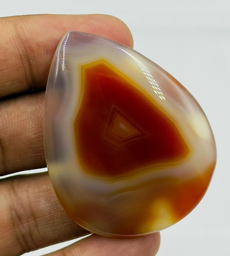 May include: A polished, teardrop-shaped agate gemstone featuring a translucent, light purple outer edge and a vibrant, deep orange-red center with distinct, concentric triangular banding patterns visible within the stone's smooth surface.