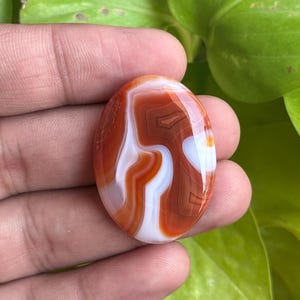 May include: An oval-shaped polished cabochon gemstone featuring distinct bands of deep burnt orange and translucent white. The stone has a smooth, glossy surface with swirling, organic patterns that create a layered appearance throughout.