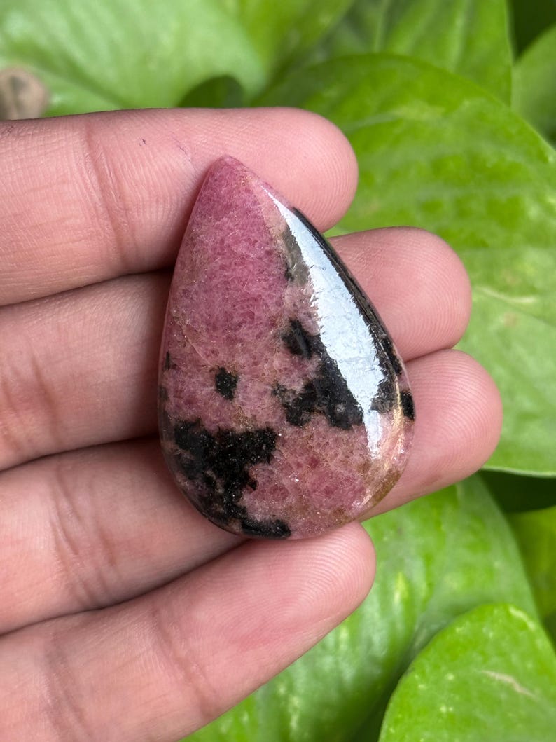 K&ouml;nnte beinhalten: Ein polierter, tropfenf&ouml;rmiger Edelstein mit einem rosa und schwarzen Muster. Der Stein hat eine gl&auml;nzende Oberfl&auml;che und wird in einer Hand vor einem Hintergrund aus gr&uuml;nem Laub gehalten. Die rosa Farbe dominiert, mit schwarzen Einschl&uuml;ssen.