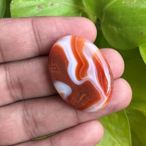 May include: An oval-shaped cabochon gemstone featuring smooth, polished surfaces with distinct, swirling bands of deep burnt orange, vibrant red, and opaque white colors, showcasing natural layered patterns throughout the stone.