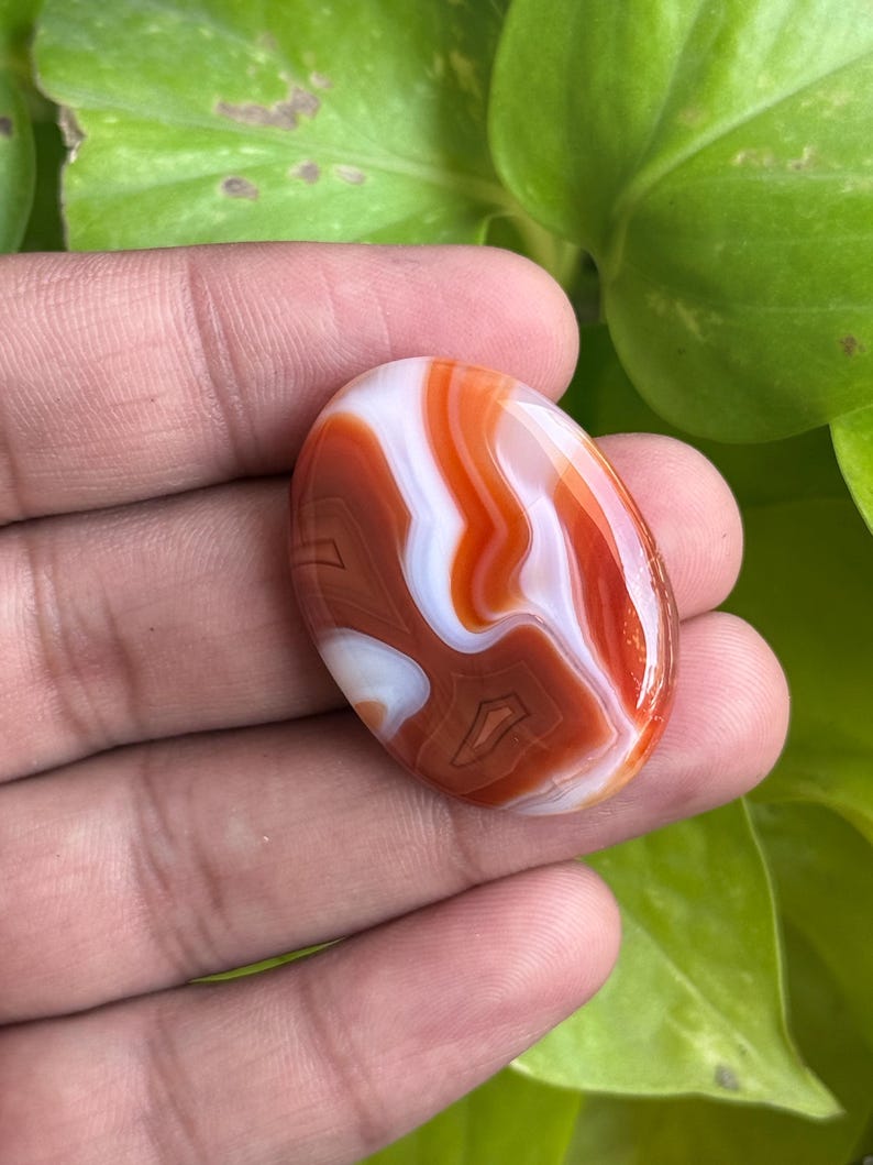 May include: An oval-shaped cabochon gemstone featuring smooth, polished surfaces with distinct, swirling bands of deep burnt orange, vibrant red, and opaque white colors, showcasing natural layered patterns throughout the stone.