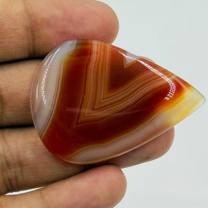 Pode incluir: Uma pedra preciosa de &aacute;gata cornalina polida em forma de l&aacute;grima, com bandas conc&ecirc;ntricas distintas de vermelho ferrugem, laranja queimado e branco transl&uacute;cido. A pedra tem uma superf&iacute;cie lisa e brilhante com reflexos de luz vis&iacute;veis.