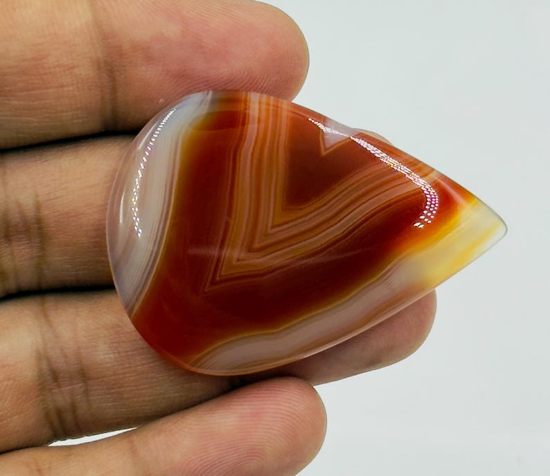 Pode incluir: Uma pedra preciosa de &aacute;gata cornalina polida em forma de l&aacute;grima, com bandas conc&ecirc;ntricas distintas de vermelho ferrugem, laranja queimado e branco transl&uacute;cido. A pedra tem uma superf&iacute;cie lisa e brilhante com reflexos de luz vis&iacute;veis.
