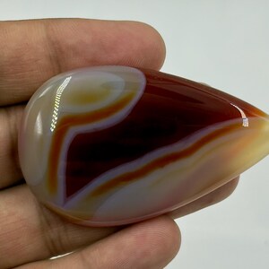 May include: A polished, teardrop-shaped agate gemstone featuring smooth, concentric bands of deep mahogany, translucent amber, and creamy white. The stone has a high-gloss finish that reflects light, highlighting its layered, organic color patterns.