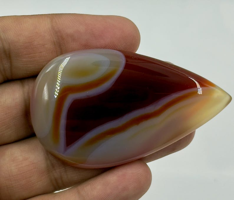 May include: A polished, teardrop-shaped agate gemstone featuring smooth, concentric bands of deep mahogany, translucent amber, and creamy white. The stone has a high-gloss finish that reflects light, highlighting its layered, organic color patterns.