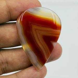Pode incluir: Uma pedra preciosa de &aacute;gata polida em forma de l&aacute;grima com faixas vibrantes de vermelho profundo, laranja queimado e branco transl&uacute;cido. A pedra exibe um padr&atilde;o distinto em forma de chevron nas suas camadas, com um acabamento de superf&iacute;cie liso e brilhante.