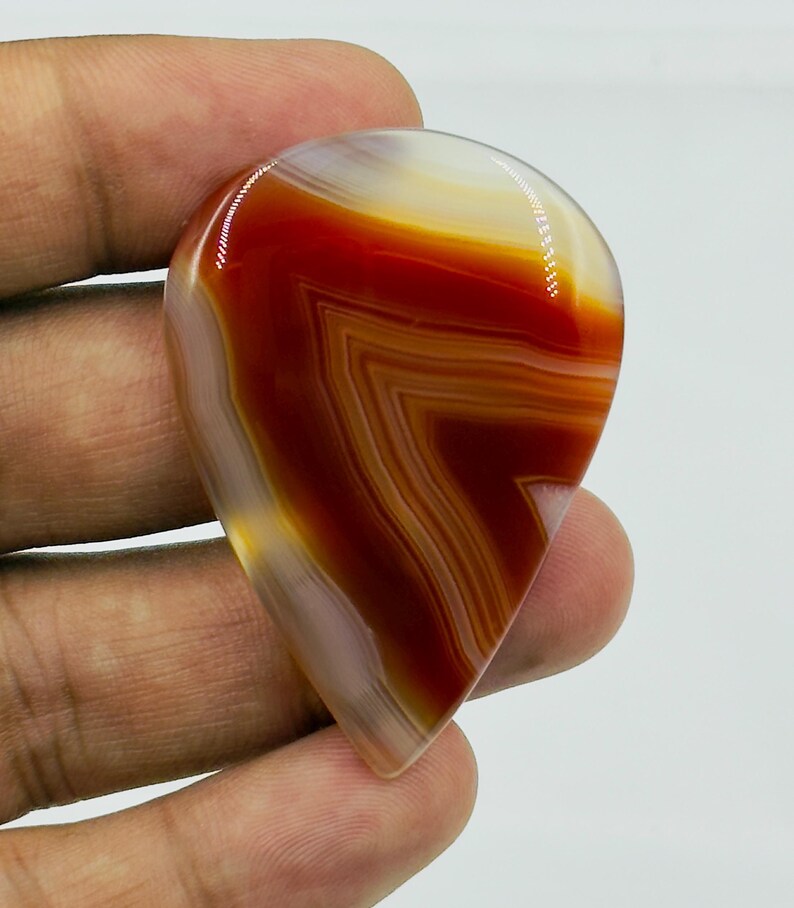 Pode incluir: Uma pedra preciosa de &aacute;gata polida em forma de l&aacute;grima com faixas vibrantes de vermelho profundo, laranja queimado e branco transl&uacute;cido. A pedra exibe um padr&atilde;o distinto em forma de chevron nas suas camadas, com um acabamento de superf&iacute;cie liso e brilhante.