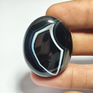 May include: A polished, oval-shaped black agate gemstone with a striking white and light blue banded pattern. The stone is held between fingers, showcasing its smooth, glossy surface. The design features a central, shield-like shape.