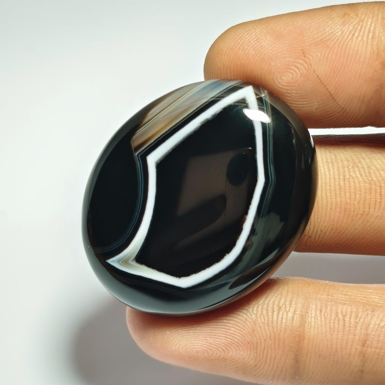 May include: A polished, oval-shaped gemstone with a striking black and white pattern. The stone features a prominent white geometric design against a black background, with bands of brown and white. The gem is held between fingers.